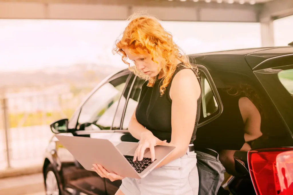 Chica buscando en su ordenador cómo importar vehículos a su país apoyando su espalda en su coche negro