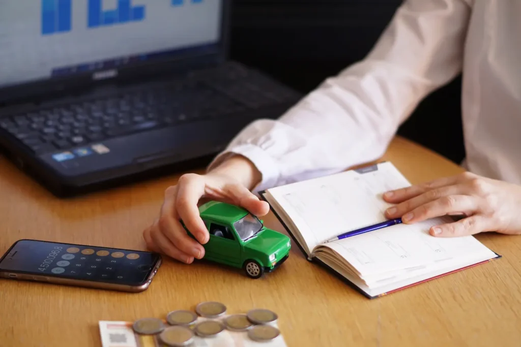 Plano medio de una persona sentada en un escritorio con un coche verde en la mano, una libreta, una calculadora y unas monedas calculando el impuesto de importación de vehículos